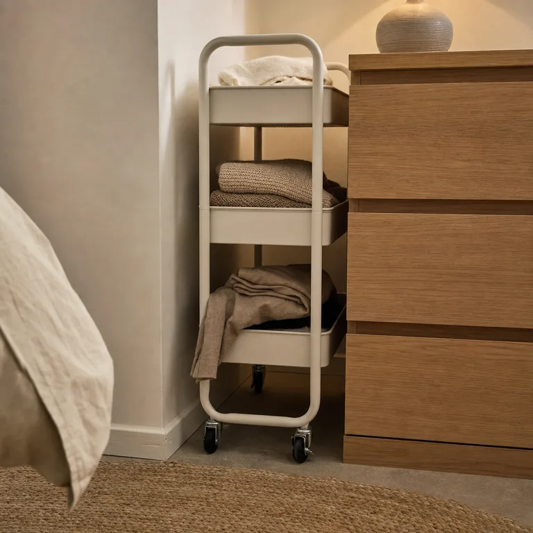 A slim rolling cart holding folded clothes beside a bedroom dresser.