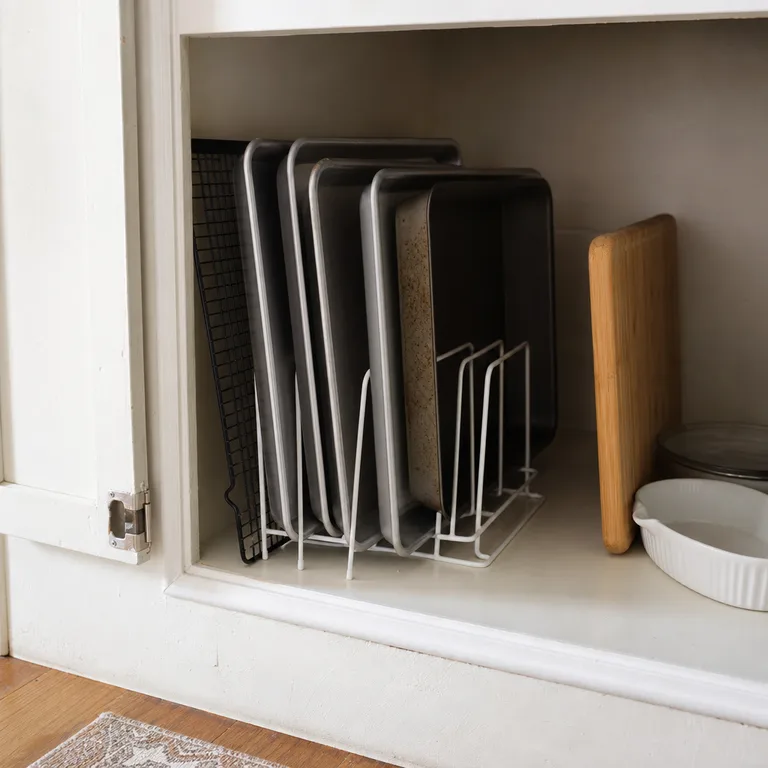 Freestanding wire rack holding baking sheets upright inside a narrow lower cabinet.